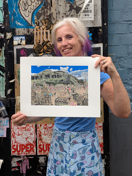 Liz holding a print of a Collage of a Scottish mountainous landscape with buildings and text, resembling a cityscape.