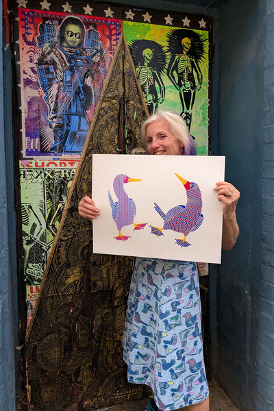 Print of a pair of dancing birds, one with pink, purple and blue with dotty feet and one in red, blue and purple with blue feet with a heart pattern held by Liz