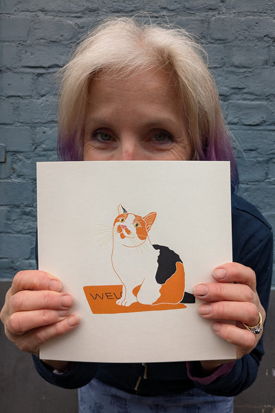 Print of a calico cat sitting on the mat held by Liz