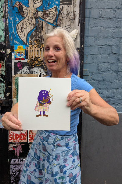 Liz holding a print of a purple haggis character in a kilt playing bagpipes