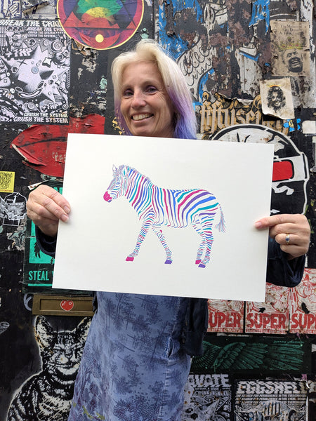 A colourful screen print of a zebra walking in profile, filled with vibrant, multicoloured zebra-stripe patterns in pink, blue, purple, and teal on a white background held by Liz
