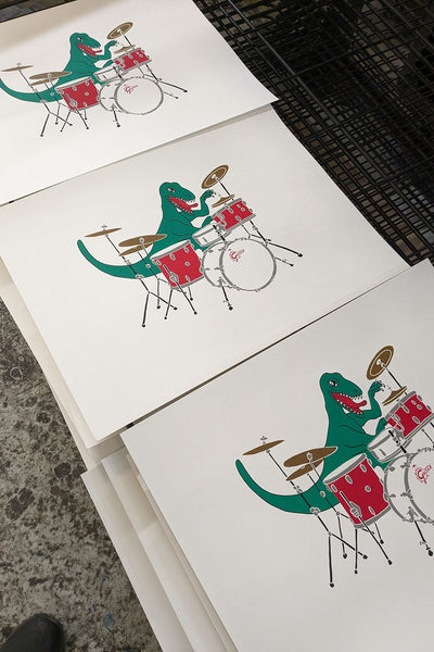 Drumming dinosaur screen print by Liz Whiteman Smith on the drying rack